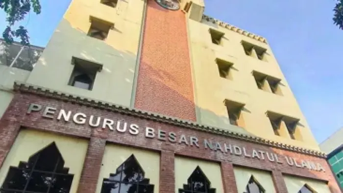 Tampak depan Gedung PBNU dari sudut bawah, dengan papan nama "Pengurus Besar Nahdlatul Ulama" terpasang di dinding bata merah. Bangunan berwarna krem itu memiliki jendela berbentuk lengkung runcing di lantai dasar dan deretan jendela persegi di lantai atas.