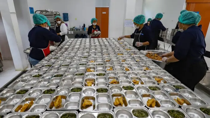 Sejumlah pekerja dapur berhairnet dan bermasker mengisi wadah alumunium berisi sayur dan lauk goreng yang tersusun di meja panjang, menunjukkan proses produksi MBG dalam skala besar.