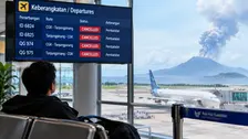 Gambar menampilkan seorang penumpang duduk membelakangi kamera sambil menatap layar informasi keberangkatan (departures) yang menunjukkan empat penerbangan berstatus "CANCELLED",