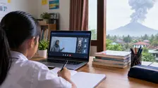 Seorang siswa mengikuti pembelajaran daring dari rumah menggunakan laptop dan buku pelajaran, dengan ilustrasi Gunung Anak Krakatau yang mengeluarkan asap di latar belakang.