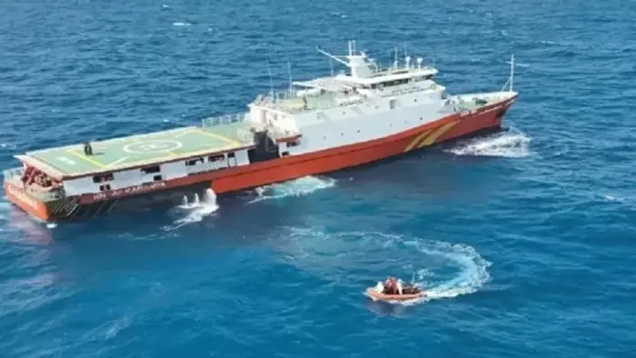 Foto udara memperlihatkan sebuah kapal berlambung merah dan berbangunan putih dengan geladak helikopter di bagian haluan, berlayar di laut biru. Di sisi depannya, satu perahu karet oranye berisi beberapa personel bergerak menjauh dari kapal induk, meninggalkan jejak ombak putih di permukaan air.