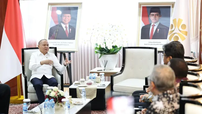 Dalam foto tampak seorang pejabat mengenakan kemeja putih sedang berbicara sambil menggerakkan kedua tangannya, duduk di kursi berwarna krem di ruang pertemuan resmi. Di latar belakang terpasang dua foto besar Presiden Jenderal TNI (Purn) H. Prabowo Subianto dan Wakil Presiden Gibran Rakabuming, diapit oleh bendera Merah Putih dan rangkaian bunga anggrek putih.