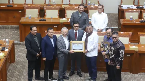 Foto memperlihatkan sesi foto bersama di ruang rapat Komisi III DPR RI, dengan latar belakang deretan meja dan kursi berwarna kayu cokelat khas ruang rapat parlemen. Delapan pria berdiri berjajar di barisan depan, sementara dua orang lainnya berdiri di barisan belakang.
