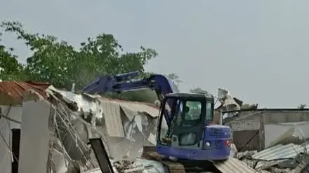 Ekskavator biru membongkar bangunan beratap seng dengan puing yang berserakan di sekitarnya. Di latar belakang tampak pepohonan dan langit berawan.
