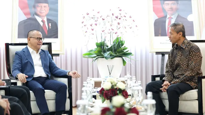 Dua pria duduk berhadapan di kursi putih berbingkai hitam sambil berbincang di ruang kerja. Pria di kiri mengenakan blazer biru dan tersenyum sambil menggerakkan tangan kanannya. Pria di kanan mengenakan batik cokelat dengan tangan terlipat di pangkuan. Di belakang mereka ada rangkaian anggrek ungu dan dua bingkai potret Presiden dan Wakil Presiden, sedangkan di meja depan ada bunga mawar merah dan putih serta botol air.
