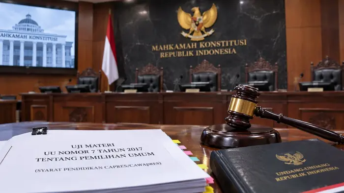 Ilustrasi ruang sidang Mahkamah Konstitusi menampilkan palu sidang, berkas permohonan uji materi UU Nomor 7 Tahun 2017 tentang Pemilihan Umum, serta buku peraturan perundang-undangan. Latar belakang memperlihatkan kursi persidangan dan lambang Garuda sebagai simbol lembaga peradilan konstitusi. Visual ini merepresentasikan pengujian ketentuan hukum mengenai syarat pendidikan capres dan cawapres yang tengah menjadi perhatian para pemohon.