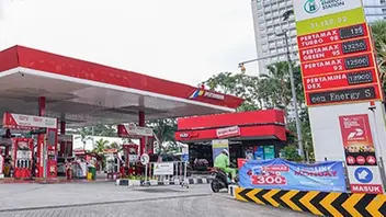 SPBU Pertamina dengan kanopi merah dan putih serta deretan dispenser BBM di bawahnya. Di sisi kanan berdiri papan harga BBM yang memuat sejumlah jenis Pertamax. Seorang pengendara sepeda motor tampak melintas di dekat pintu masuk, dengan gedung bertingkat dan pepohonan di latar belakang.