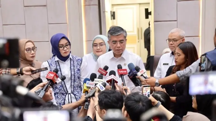 Seorang pria berkemeja putih dengan pin di dada berbicara di depan puluhan mikrofon dan ponsel milik awak media yang mengerumuninya. Di sisinya berdiri dua perempuan berhijab, dan di belakangnya seorang pria berkacamata mengenakan kemeja putih. Terlihat mikrofon dari sejumlah media televisi, di antaranya CNN, tvOne, dan Metro TV. Latar belakangnya dinding krem dengan pintu di bagian tengah.
