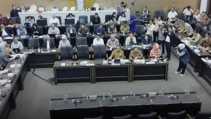 Suasana ruang rapat dengan meja-meja berbentuk huruf U yang dipenuhi peserta rapat. Beberapa juru foto tampak mengambil gambar, dengan ornamen emas berlatar biru dan kuning di dinding belakang.