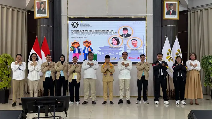 Sebelas orang berdiri berjajar di panggung dengan kedua tangan disilangkan di dada. Sebagian mengenakan kemeja putih, sebagian jaket krem atau hitam, dan seorang di tengah berpeci hitam memakai selendang kuning. Di belakang mereka terdapat layar bertulisan "Pembekalan Motivasi Pemberangkatan Calon Pekerja Migran Indonesia Penempatan ke Jerman dan Korea Selatan", dengan bendera Merah Putih dan foto presiden serta wakil presiden di sisi kiri dan kanan.