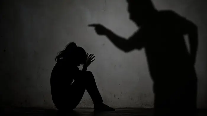 Siluet seorang perempuan duduk meringkuk dengan gestur ketakutan di sudut ruangan, sementara bayangan sosok lain tampak mengintimidasi dari kejauhan