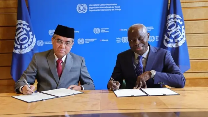 Dua pejabat duduk berhadapan di meja kayu dengan latar belakang backdrop biru bertuliskan International Labour Organization dalam tiga bahasa.