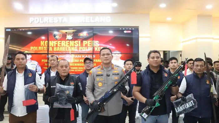 Kasat Reskrim Polresta Barelang Kompol Agung Tri Poerbowo (kanan) memegang senjata laras panjang berwarna hijau loreng. Di sebelah kiri, Kapolresta Barelang memegang senjata laras panjang berwarna hitam dan biru. Keduanya mengenakan sarung tangan hitam. Di latar belakang tampak layar konferensi pers Polresta Barelang.