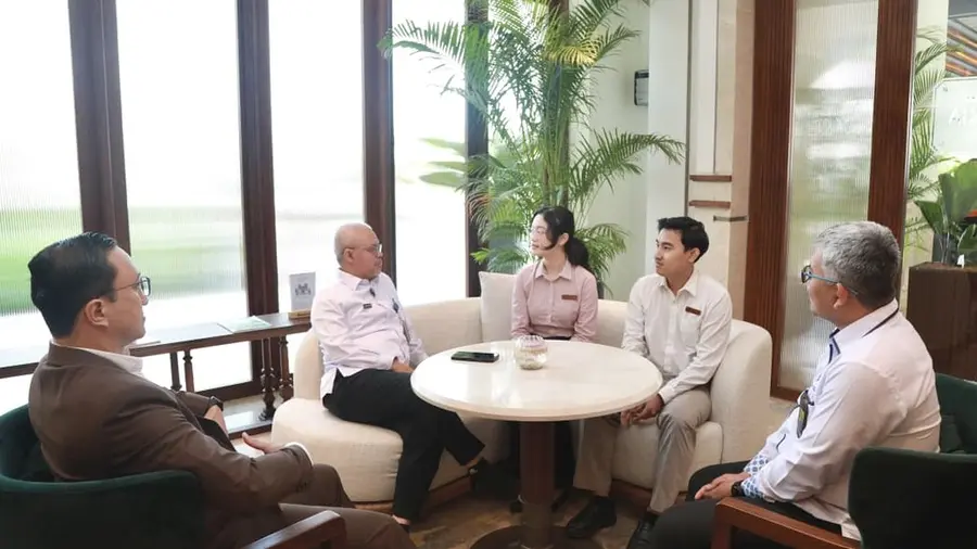 Kemnaker Manfaatkan AI untuk Verifikasi Mitra dan Peserta MagangHub Lima orang duduk mengelilingi meja bundar putih di ruang lobi hotel. Dua orang berseragam duduk di sofa krem, sementara tiga orang lainnya duduk di kursi hijau di sisi kiri dan kanan. Di latar belakang tampak jendela besar dan tanaman palem.