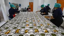 Sejumlah pekerja dapur berhairnet dan bermasker mengisi wadah alumunium berisi sayur dan lauk goreng yang tersusun di meja panjang, menunjukkan proses produksi MBG dalam skala besar.