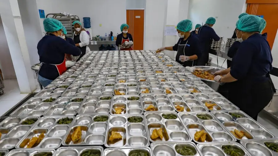 62 SPPG di Banten Terancam Ditutup Permanen Jika Tak Urus SLHS Sejumlah pekerja dapur berhairnet dan bermasker mengisi wadah alumunium berisi sayur dan lauk goreng yang tersusun di meja panjang, menunjukkan proses produksi MBG dalam skala besar.