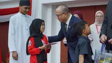 Menteri P2MI Mukhtarudin, mengenakan jas biru tua dan kacamata, tampak menyerahkan amplop bantuan pendidikan kepada seorang anak perempuan berjilbab hitam berseragam merah. Anak tersebut menerima amplop dengan kedua tangan sambil menatap ke arah Mukhtarudin
