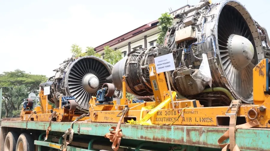 Polda Kepri Ungkap Impor Ilegal Tiga Mesin Pesawat Bekas dari Singapura Senilai Rp30 Miliar Mesin pesawat berukuran besar diletakkan di atas bak truk trailer berwarna hijau dengan rangka penyangga oranye. Di dekatnya terpasang papan bertulisan "Barang Bukti: Mesin Pesawat/Engine Turbo" dan pita kuning pembatas. Di latar belakang tampak gedung bertingkat dan pepohonan.