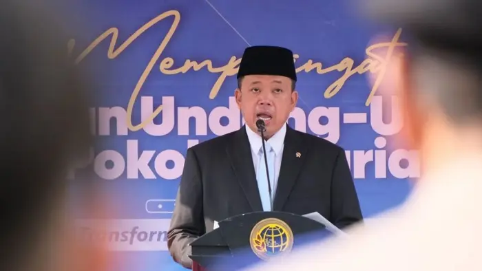 Seorang pria berpeci hitam, berjas gelap, dan berkemeja putih dengan dasi biru muda berbicara di balik podium. Di depan podium terpasang lambang berbentuk bola dunia berwarna emas. Di belakangnya terpasang spanduk biru bertuliskan "Memperingati" dan sebagian kalimat yang tampaknya memuat "Undang-Undang Pokok Agraria". Sosok di latar depan tampak buram.