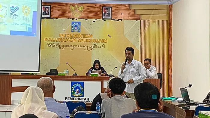 Seorang pria berkemeja putih berdiri sambil memegang mikrofon saat menjelaskan materi di hadapan warga yang duduk di barisan depan. Di belakangnya, dua peserta lain duduk di meja panel berlatar tulisan "Pemerintah Kalurahan Wukirsari", sementara layar proyektor di sisi kiri menampilkan materi dasar hukum pelindungan pekerja migran.