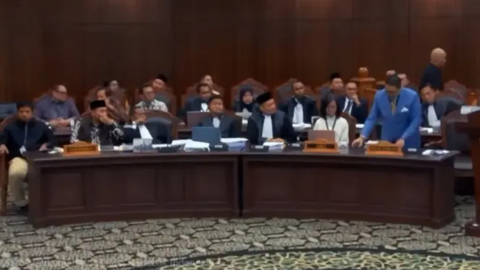Tangkapan layar ruang sidang dengan para peserta sidang duduk di balik meja panjang berwarna cokelat tua. Seorang pria berjas biru berdiri di dekat podium di sisi kanan, sementara yang lain duduk menghadap ke depan. Dinding kayu bernuansa cokelat dan lambang Garuda di podium tampak di latar belakang.