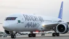 Pesawat berbadan lebar warna putih bertuliskan "Riyadh Air" dalam huruf Latin dan Arab di badan pesawat. Sirip ekornya berwarna biru dan terdapat bendera Arab Saudi di dekat pintu depan. Pesawat berada di landasan bandara dengan kendaraan pendukung di belakangnya.