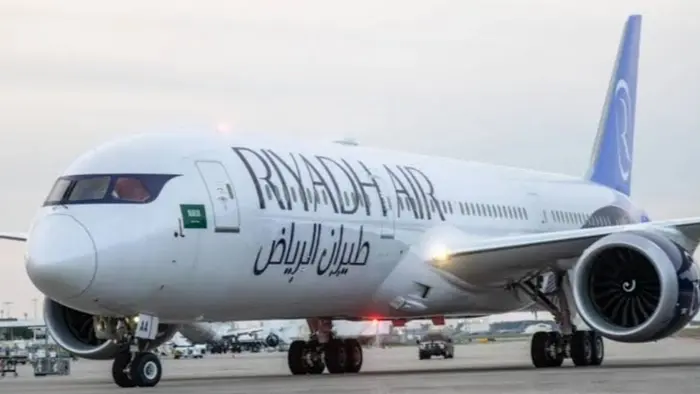 Pesawat berbadan lebar warna putih bertuliskan "Riyadh Air" dalam huruf Latin dan Arab di badan pesawat. Sirip ekornya berwarna biru dan terdapat bendera Arab Saudi di dekat pintu depan. Pesawat berada di landasan bandara dengan kendaraan pendukung di belakangnya.