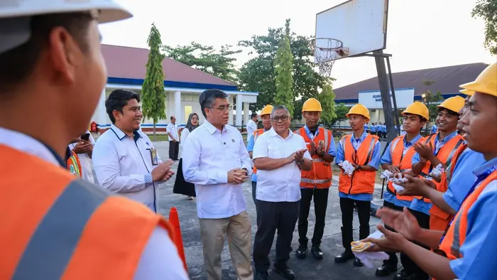 Sejumlah peserta pelatihan berhelm kuning dan berompi oranye berdiri melingkar sambil bertepuk tangan di lapangan terbuka. Dua pria berkemeja putih berdiri di tengah lingkaran, ditemani seorang pria berkemeja putih lain di sisi kiri. Di latar belakang tampak ring basket, pohon, dan gedung berbumbung merah.
