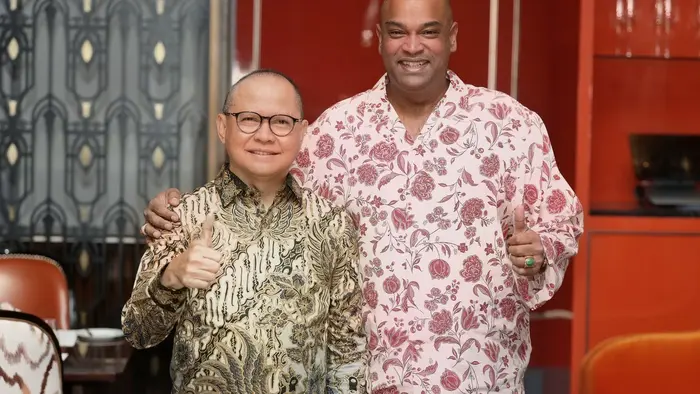 Dua pria berdiri berdampingan sambil tersenyum dan mengacungkan jempol. Pria di sebelah kiri berkacamata dan mengenakan batik cokelat keemasan. Pria di sebelah kanan berbaju bermotif bunga merah muda dan merangkul bahu pria di sebelahnya. Latar belakangnya ruang makan bernuansa merah dan oranye.