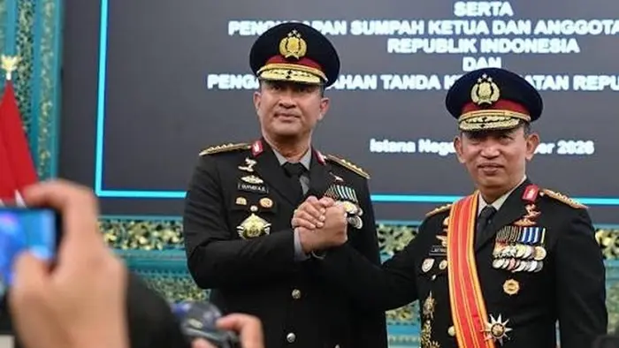 Dua perwira polisi berseragam dinas hitam dengan topi baret dan tanda pangkat berdiri berdampingan sambil berjabat tangan. Orang di sebelah kanan mengenakan selempang kuning-merah dan sejumlah tanda jasa di dadanya. Di latar belakang tampak layar bertulisan acara pelantikan dan penganugerahan tanda kehormatan di Istana Negara.