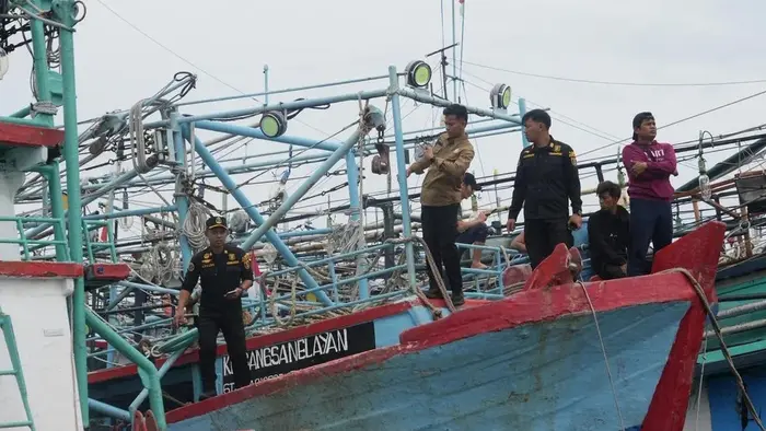 Beberapa petugas berseragam hitam dan cokelat berdiri di dek sebuah kapal perikanan berlambung biru dan merah. Di belakang mereka tampak tiang, lampu sorot, dan tali yang menumpuk, serta beberapa orang berpakaian biasa duduk dan berdiri di dekatnya. Kapal lain bersandar di sekitarnya, dengan langit berawan.