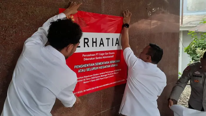 Dua petugas berkemeja putih menempelkan papan segel merah bertuliskan "PERHATIAN" di dinding granit kantor PT Ceger Sari Buana. Segel itu memuat keterangan bahwa perusahaan dikenai sanksi administratif penghentian sementara sebagian atau seluruh kegiatan usaha berdasarkan Keputusan Direktur Jenderal Pelindungan Nomor 44 Tahun 2026. Seorang anggota Polri berseragam coklat turut mendampingi proses pemasangan.