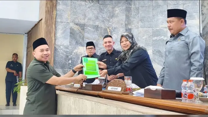 Seorang pria berpeci hitam dan berkemeja hijau army menyerahkan map hijau berlogo PKB kepada seorang perempuan berkerudung motif yang duduk di meja pimpinan sidang. Di belakang mereka, dua pria berseragam dinas berdiri sambil tersenyum, sementara satu pria berpeci dan berkemeja abu-abu berdiri di sisi kanan. Di atas meja terdapat papan nama, botol air mineral, dan gelas.