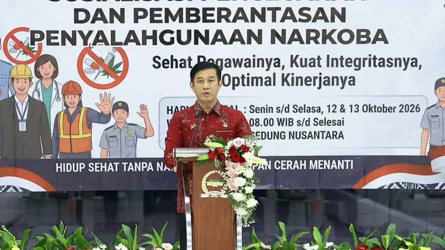 Setjen DPR Bentuk Tim Khusus Deteksi Dini Narkoba Seorang pria berbatik merah berbicara di balik podium berlambang Setjen DPR RI yang dihias rangkaian bunga mawar putih dan merah muda. Di belakangnya terbentang spanduk bertuliskan "Sosialisasi Pencegahan dan Pemberantasan Penyalahgunaan Narkoba" dengan slogan "Sehat Pegawainya, Kuat Integritasnya, Optimal Kinerjanya", lengkap dengan ilustrasi pekerja dan simbol larangan narkoba. Tanaman hijau tertata di bagian depan panggung.