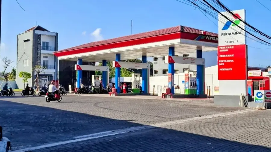 Pemkab Gianyar Tetapkan WFH Bagi PNS Imbas Antrean Panjang BBM SPBU Pertamina dengan kanopi beratap genteng merah dan tiang biru, berjajar tiga pulau dispenser. Papan harga di sisi kanan memuat jenis BBM Pertamax Turbo, Pertamax, Pertamina Dex, dan Pertalite. Beberapa pengendara sepeda motor melintas di jalan paving, dengan petugas dan kendaraan tampak di area pengisian serta langit cerah di latar belakang.